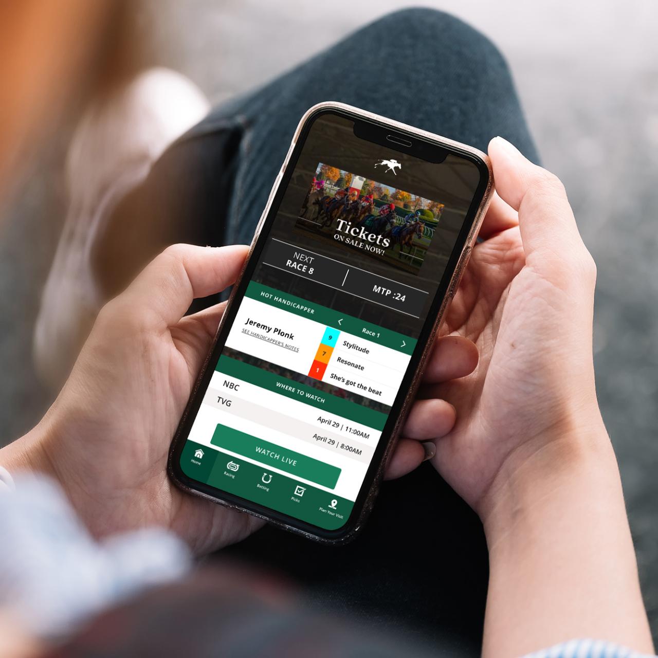 Hands holding a phone showing Keeneland App-Home screen. This new feature has the hottest handicappers, watch live, and your favorites horse picks per race. 